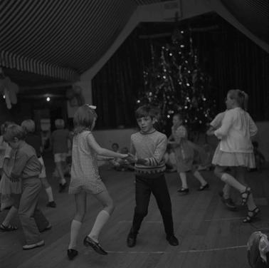 Two children dancing in a hall in front of a Cristmas tree.  Other children in the background also dancing.