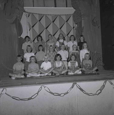 Eighteen school children llined up in three rows sitting, kneeling and standing on a stage.