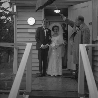 A bride and groom at the door of a weatherboard church.  A man is holding an umbrella over the couple