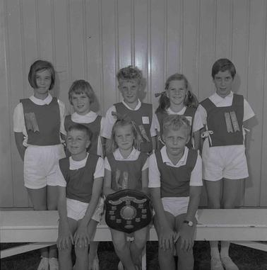 Eight girls and boys in school sports uniform standing in two rows.  A girl in front row is holding a trophy board.
