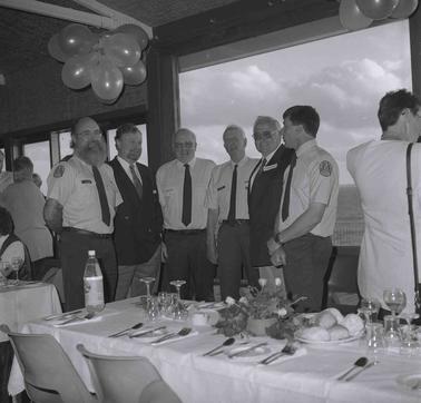 Seven men in white shirts and ties (one with a coat on) posing for a photograph around the corner of a table set for a dinner.  A buch of balloons above,