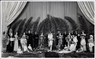 This image shows a group of twenty-four men, women and children standing and sitting in a theatre.  The women are dressed in long formal dressed with crowns on their heads and large bouquets of flowers.  The woman at the centre of the group is holding a sceptre.  One man is dressed as a judge, another is in a suit and the remainder of the men are dressed as ministers of the church.  A pageboy is holding a cushion on the far right-hand side.  Large palm fronds are standing behind the group as well on the floor at the front.  Large curtains are at the back and sides of the theatre.  