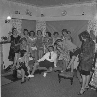 A group of thirteen women in a jovial mood dressed in ‘1920s’ costumes, some standing and three seated posing for a photograph. 