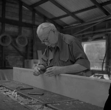 A mature man wearing rolled up shirt sleeves at a woodworking bench with a timber plank.