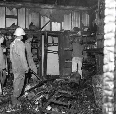 A burned out interior being inspected by three men (one wearing a safety helmet) and two adolescent boys looking on.