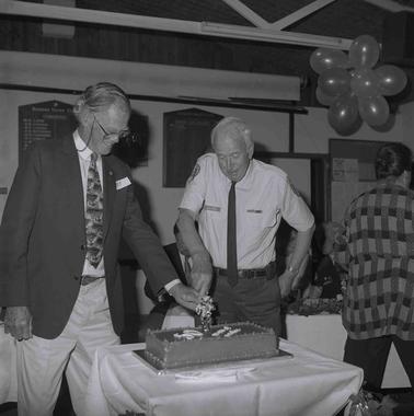 two men at a white cloth covered table cutting a cake together.
