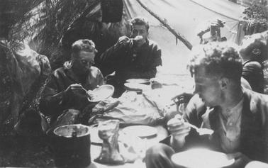 Camping scene. Close up of four young men seated cross-legged around mat spread on ground and eating from enamel plates. Billy can left foregrouno date. Tent in backgrouno date.