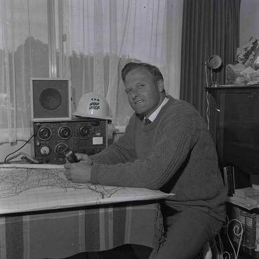 A man with a radio at his elbow seated at a table under a window .