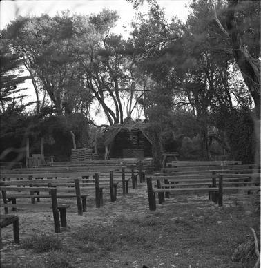 Outdoor chapel setting with row of wooden benches on a grassy glade facing a brushwood ‘grotto’ altar.