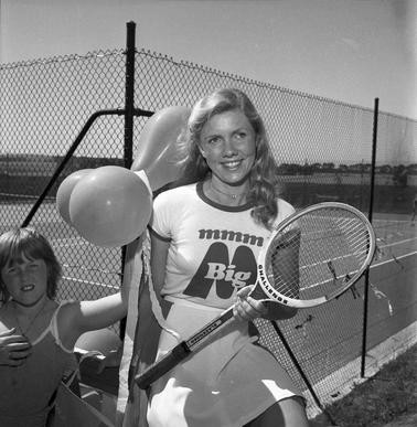 A girl with a tennnnis racquet standing at the outside corner of a tennis court with a young boy and a balloon.