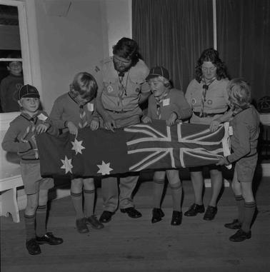 Four boys inCub uniforms inspecting an Australian flag while one male and one female Cub Leader look on.