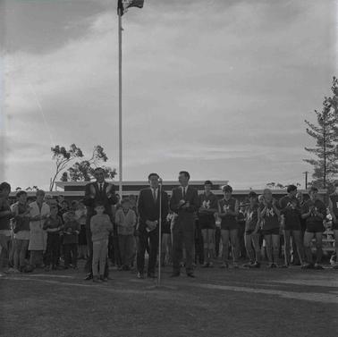 Three men at a microphone and a fllying flag with footballers and others in front of a building.