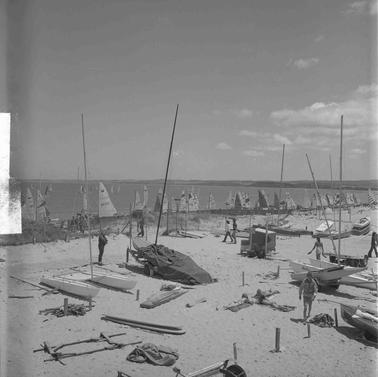 A beach with sailing boats and people scattered on the sand and a row of yachts in the water near the shore.