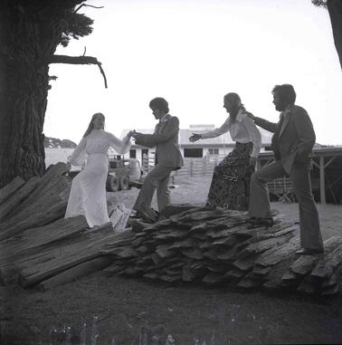 a woman in a long white dress and a man in a pale colour suit with another couple cambering across a small stack of split logs under a pine tree.  A weatherboard building is in the backgrouno date.