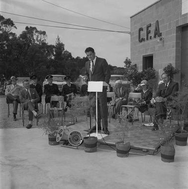 One man on an outdoor podium speaking into a microphone while eight men and one woman are seated behind him in front of the Balnarring CFA building.