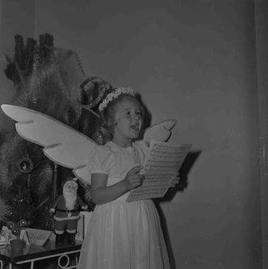 A girl standing in front of a Christmas tree, dressed as an angel singing, holding a sheet of music.