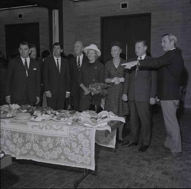Six men and two ladies standing in a row beside a festive table laden with food.  The womanin the centre is holding a flower bouquet.