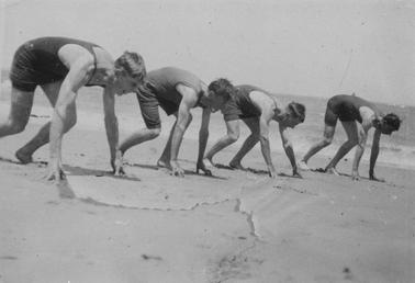 Four young men, lined up in starting crouch for a race. Wearing 20’s style one piece bathing suits. On beach, sea in backgrouno date.