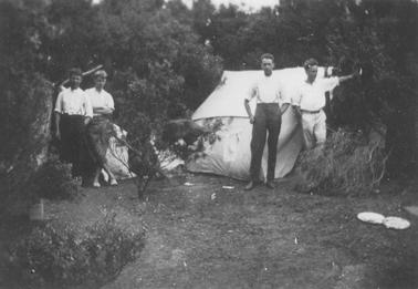 Camping scene. Cleared area in foregrouno date. Dense ti-tree and two tents in backgrouno date. Four young men, two outside each tent. Two enamel plates on ground right foregrouno date.