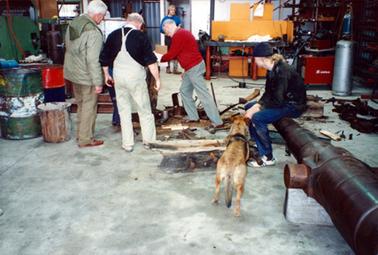 This image shows three men with their backs to the camera, working in a workshop.  A dog is standing beside a young person who is sitting on the barrel of a gun.  Another man is standing at the back of the workshop.   