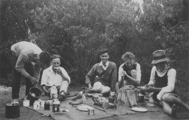 Five young people dining outdoors. Man on left side is bending forward, pouring liquid from a billy into enamel mugs. Next to him is young male in white shirt eating; then male in sweater and shorts looking at the camera. Fourth figure has short curly hair and glasses, and is cutting something on tree stump. Fifth figure has shorts, dark top, white shirt and man’s hat, and is holding a plate. Picnic rugs spread on grouno date. Tent and bushes in backgrouno date.