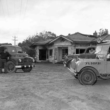Two fire trucks parked close to a weatherboard building with product logos ‘’Kodak”, Cottees’ and ‘Coka Cola’ around the door.  Two trucks and a Land Rover parked in the foregrouno date. One man leaning on a truck, another sitting inside a truck and two women near the building talking to each other.