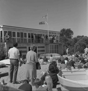 People lounging on a beach among small boat hulls while officials address them from the Yacht Club verandah.