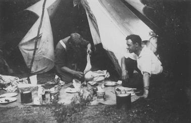 Merricks Beach. Two young men in shirt sleeves squatting in front of a tent. Picnic meal spread on ground sheet in foregrouno date. One man looking down other in profile.