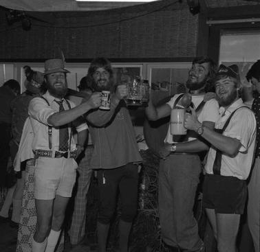 Four men, two dressed in imitation German lederhosen costume, holding up beer steines.