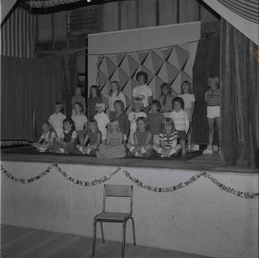 A group of twenty children forming a tableau in three rows, sitting, kneeling and standing on a stage.