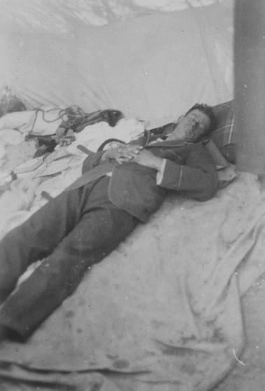 Young man in dark pajamas lying asleep on bunk inside a tent. Hands clasped across stomach. Assorted paraphernalia on left hand side.