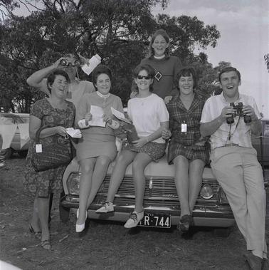 Five women and two men near a car or sitting on the car’s bonnet.