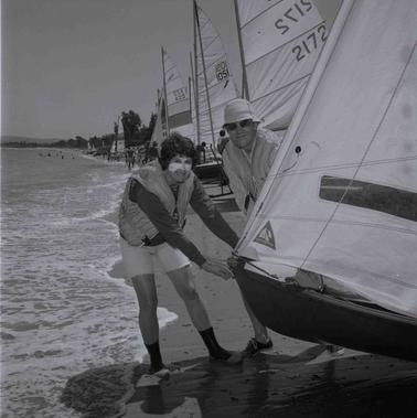 a man and woman pulling a small yacht into water off a sandy beach.  Other yachts in the backgrouno date.