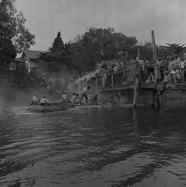 A dinghy and a raft carrying yourn men approaching a light bridge filled by spectators.