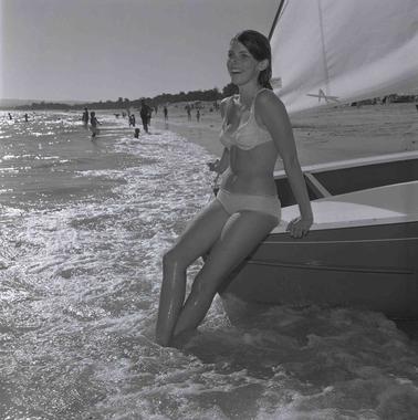 A young woman wearing a bikini leaning on the hull of a small yacht at water’s edge.  A beach and people are in the backgrouno date.