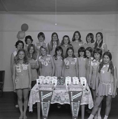 A group of seventeen girls wearing Netball uniforms, posing for a photo in two rows  Two ‘Premiers’ pennants and two celebratory cakes on a table in front.