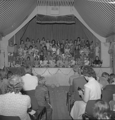 A tableau of around sixty chool children in four rows on a stage in front of an audience.  The children are singing. 