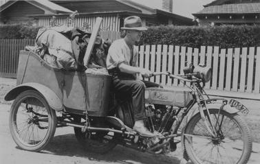 Man on motor cycle with four-wheeled carrier laden with gear behino date. Man wearing trousers, shirt with sleeves rolled above elbows, hat and light shoes. Suburban houses and fences in backgrouno date.