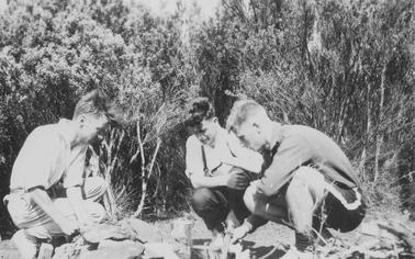 Camping scene. Three young men casually dressed, squatting around camp fire encircled by stones. Dense ti-tree in backgrouno date.