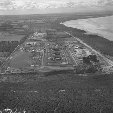 Aerial photograph of a coast with an industrial tank farm site, some tanks under construction and two compete.  A rail track in centre.