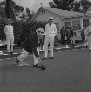 Three men at a bowling club event with others in the backgrouno date.  A clubhouse is behino date.