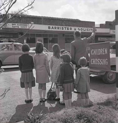 Five children waitng to cross a road with shops in the backgrouno date.