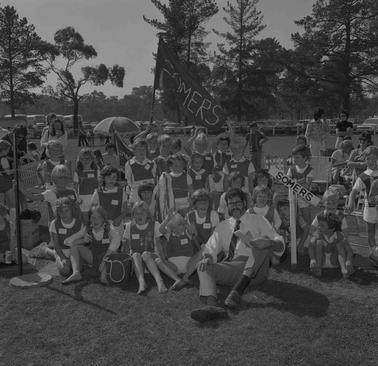 An informal group of school children dressed in sports uniforms, sitting, kneeling or standing on grass near a wire fence with an adult bearded man dressed in white shirt and tie in front row.