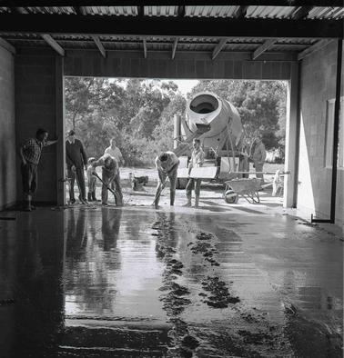 A view from the inside of a building out through an open garage door to a concrete mixing truck and a laden wheelbarrow.  Three men working the concrete with shovels.  One man is holding a wide plank, another three men and a small boy are looking on.