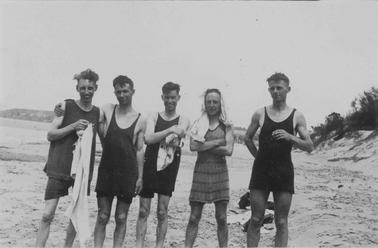 Merricks Beach, 5 young men lined up on beach, wearing singlet-style bathing suits with shirts. Two are holding towels, one has a towel draped over his head. Beach, dunes and headlands in backgrouno date.