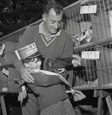 A man and girl holding a rooster in fron to chicken cages.