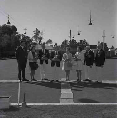 Eight men and womenon a grassy bowling club court, some with sports trophies, posing for a group photograph.