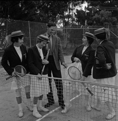 On a tennis court four our women in tennis clothing are talking to a man in coat and tie.