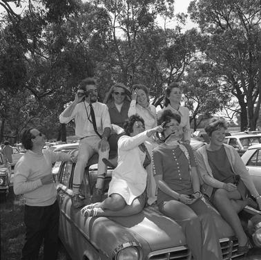 Six women and a man sitting on the bonnet and roof of a car.  One man is standing by the car.
