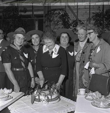 Three Girl Guides and three women: one woman is cutting a celebratory cake.